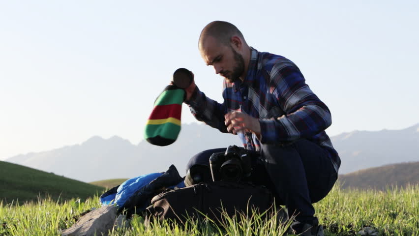 Photographer sitting and changing lenses on professional camera outdoor, with mountain landscape on the background. Traveler using a cap such as bag for photo equipment.