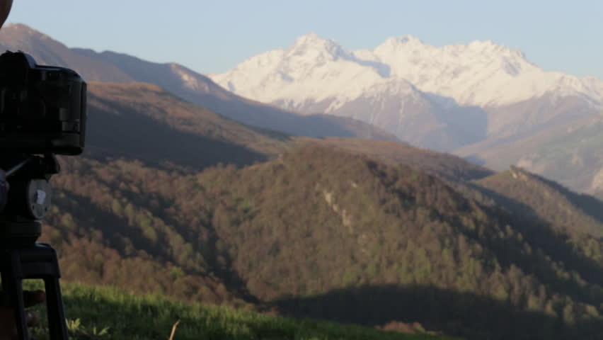 Photographer dressed as lumberjack style shooting with camera and tripod on the top of the hill. Beautiful mouintain landscape on the background, rear view.