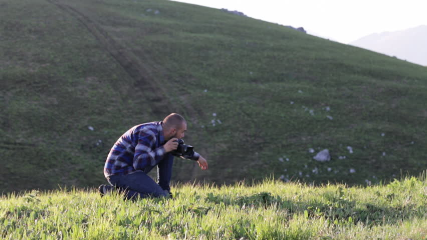Traveler standing on one knee and taking photo via professional digital camera at mountain landscape. Photographer dressed as lumberjack style.