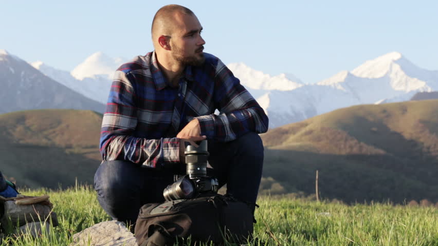 Photographer sitting and changing lenses on professional camera outdoor, with mountain landscape on the background. Traveler with photo camera dressed as lumberjack style.