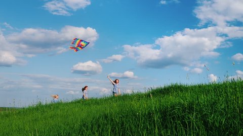 Carefree children play with a kite. Running along the green hill against the blue sky with white clouds. Happy childhood