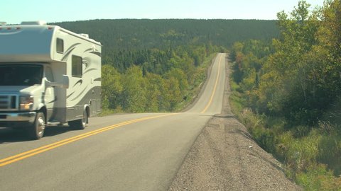 CLOSE UP: White camper car driving along empty highway in lush dense mixed forest covering the hills in the Canadian wilderness on sunny day. People on road trip in remote area in Nova Scotia, Canada