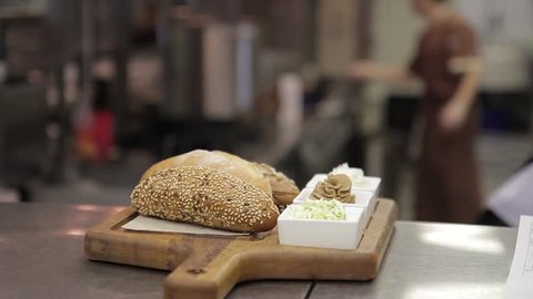 On a wooden tray lie buns with sesame and plates with condiments. In a large restaurant kitchen is ready to take out a plate with bread and different sauces and mustard. In the background, employees