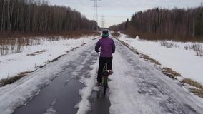 Back of woman on bicycle riding on road with thawing snow in spring park, shooting from moving near bicycle, mobile phone video.