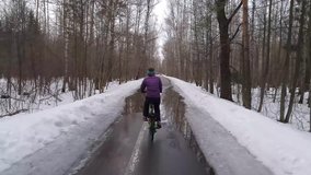 Back of woman on bicycle riding puddle on road in spring park with thawing snow, shooting from moving near bicycle, mobile phone video.