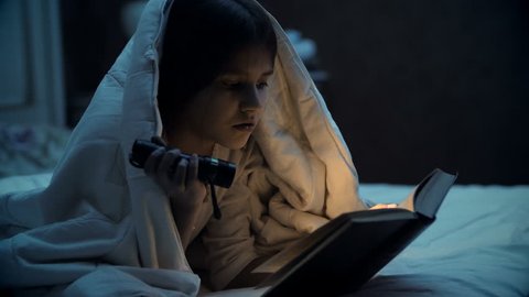 Young girl reading under blanket and lighting book with flashlight