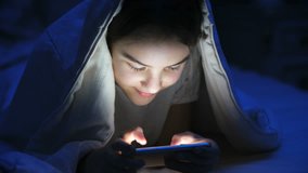 Portrait of teenage girl playing games on smartphone under blanket at night