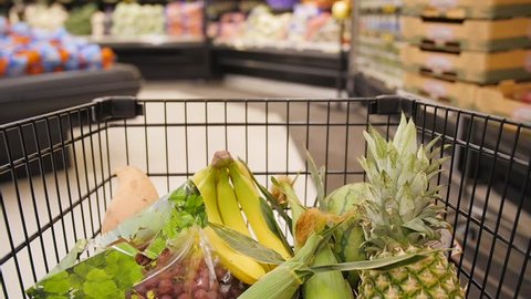 Slow-motion clip of a half-filled shopping cart moving through the produce aisle. Inside the cart, several kinds of fruits such as bananas, pineapple, watermelon, and grapes can be seen.