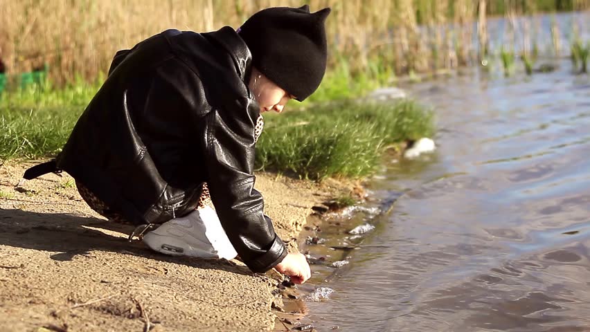 Adorable little girl wearing a black jacket playing at a river shore throwing tree branches into the water on a hot sunny spring day.