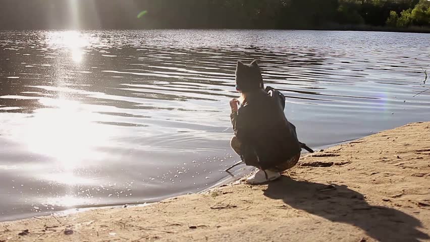 Adorable little girl wearing a black jacket playing at a river shore throwing tree branches into the water on a hot sunny spring day.