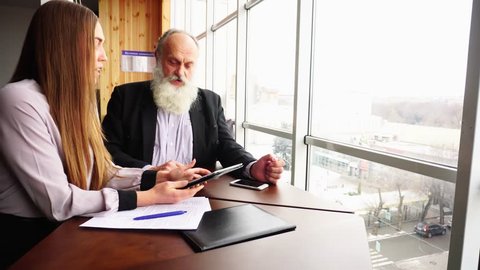 Old Retired Man Using Gadget Tablet for Dispute and Debate. Young Woman Discussing Problems in Business Matters in a Cafe in Spring. Beautiful Stately Grey Head Man With Long Beard and Young Woman.