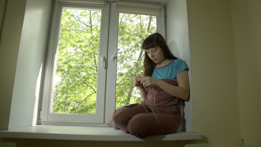 Young beautiful happy woman knits from blue yarn with knitting needles and uses smartphone sitting on window sill at bright spring day outside window trees with green leaves.