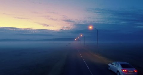Lone car on remote highway road at twilight, aerial perspective with fog landscape in background. 4K UHD