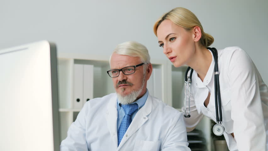 Caucasian midle aged male doctor in glasses showing and explaining something to his female colleague on a computer. Indoor.