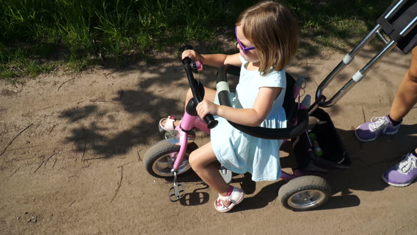 Little child walking around the village. The girl is riding a tricycle. A series of footage, batch. Sunny summer day. The girl has funny sunglasses.