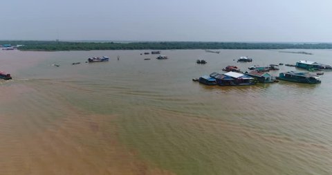 Drone footage of the floating houses where the Kam Flung Poo river is flowing into the Tonle Sap lake. The camera is going sideways in front of houses floating on the lake. A local boat is passing by.