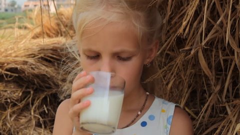 Girl drinks cow's milk from a glass against the backdrop of a haystack on a farm. The child drinks fresh milk in the summer in the village.