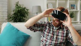 Young hipster man in VR headset watching the 360 video at home while relaxing on sofa in the living room than removing it and smiling on camera. Close up.