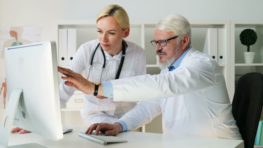 Two caucasian serious male and female doctors at medicine office talking about something looked on computer. Indoor.