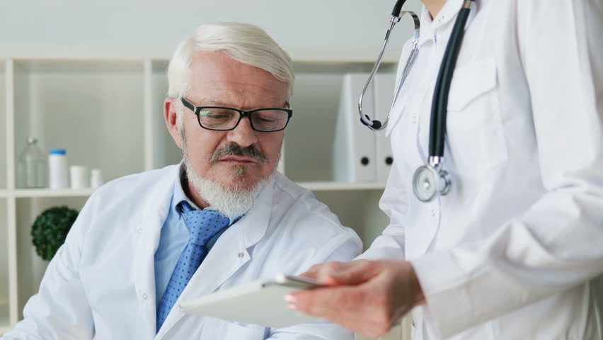 Caucasian midle aged male doctor sitting in his office and young female doctor standing in office using tablet and talking about something. Indoor.