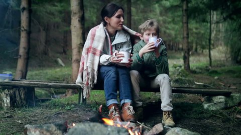 Mother with son singing and tea drinking near near forest campfire