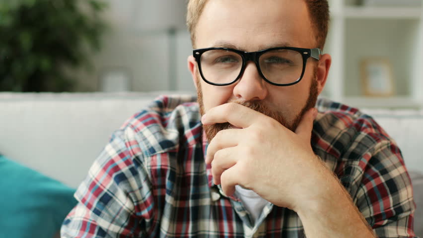 Close up portrait of young serious man in glasess sitting in front of the camera, watching film at home in the living room.
