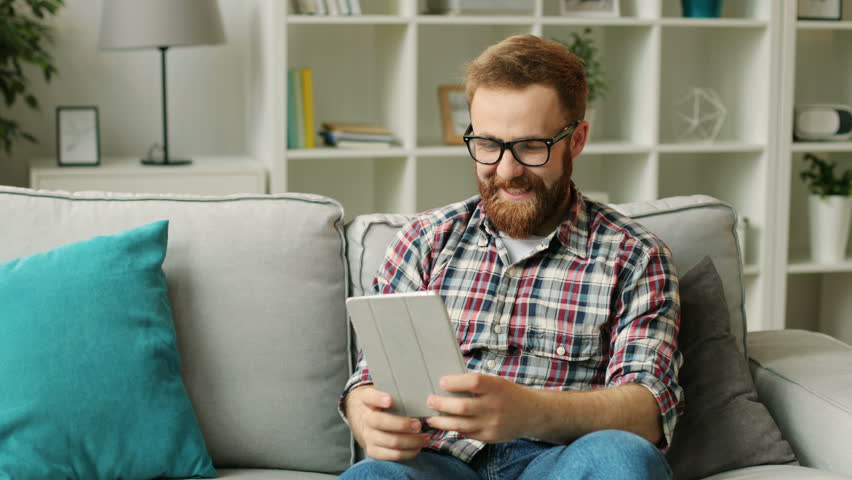 Happy attractive man having a video chat with friends on his tablet while sitting on sofa in the living room.