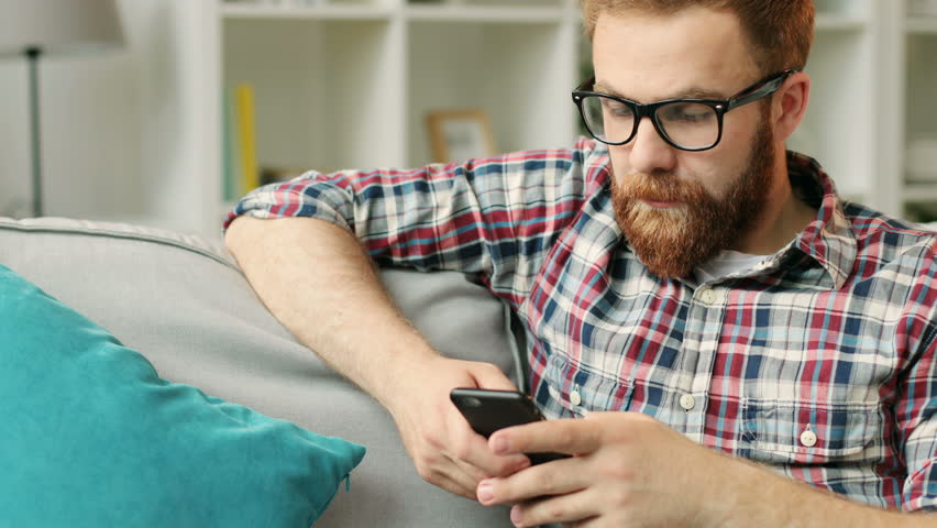 Attractive man with smartphone sitting on sofa at home in the living room and chatting with friends.