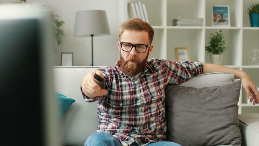 Portrait of young man in glasess sitting on a sofa turn on the tv and start watching film at home in the living room.