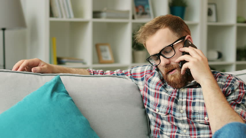 Attractive man in the glasess talking with friend on smart phone while relaxing on sofa in the living room.