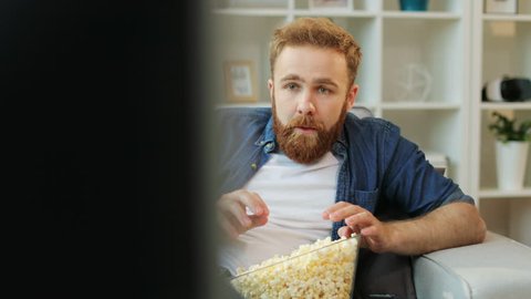 Emotional man with beard watching stressful football match, eating popcorn while sitting in the living room.