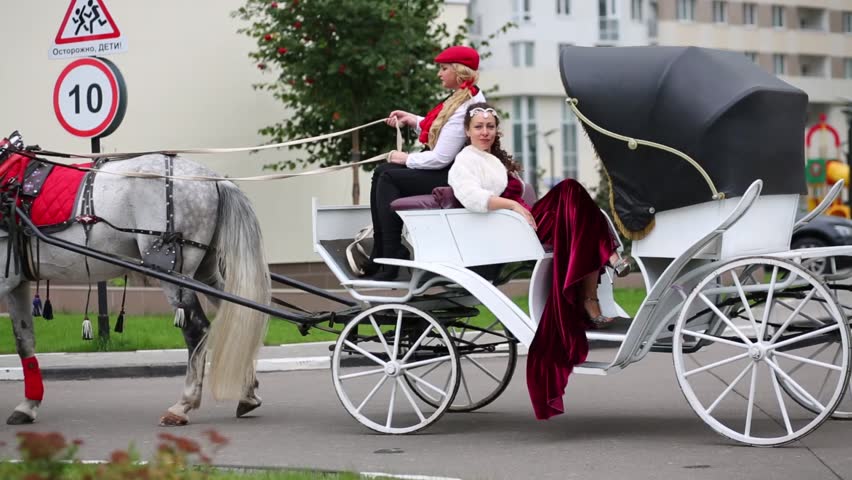 Two women are sitting at the white carriage and riding with coachman in a black coat near sign Warning! Children!.