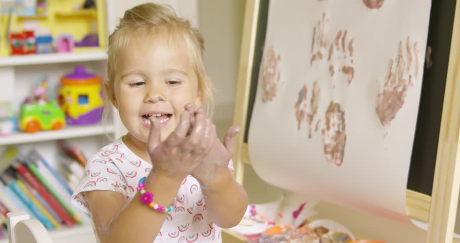 Happy attractive little blond girl artist rubbing her ands with a cute smile after applying palm prints to her abstract painting on an easel black board at home or in kindergarten.