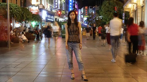 SHANGHAI JUNE 2017: Woman stands at Shanghai Nanjing East road in night timelapse. Evening time-lapse of still Asian girl standing on busy pedestrian street in with people and trams moving past.