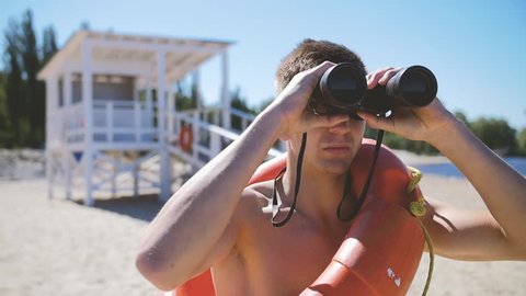 A beach lifeguard with a bare torso looks through binoculars watching suspicious personalities.In Slow Motion/