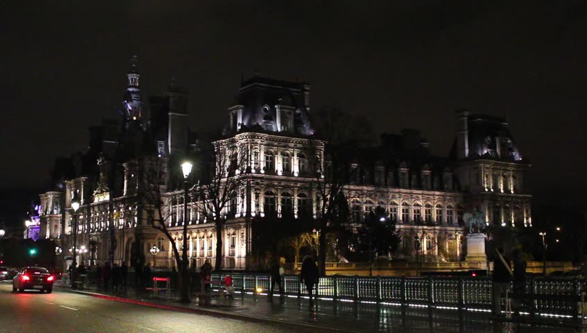 People walk on bridge on Seine river in Paris. Hotel de Ville is in the background illuminated. Winter night.
