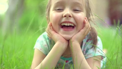 Portrait of a Happy smiling little girl lying on green grass. Cute four years old child enjoying nature outdoors. Healthy carefree kid playing outside in summer park or on backyard. 4K slow motion