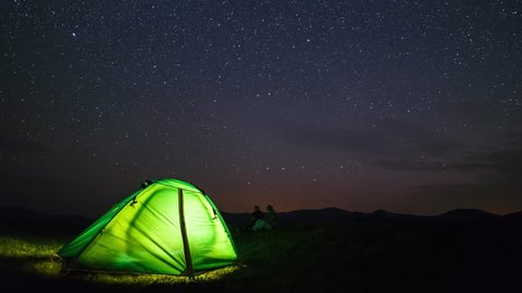 Two travelers watching the meteor shower in summer time. Timelapse of stars moving in night sky over the camp tent.