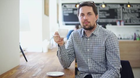 Portrait of a young man with a beard who holds a cup with hot tea or fragrant coffee in a small city cafe, the person sits at a wooden table inside