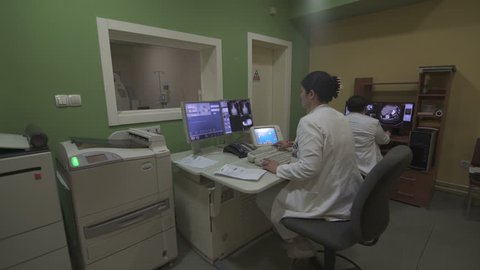 Females radiology technician and radiologist during scanning work process at control room, male patient lying on the CT scanner behind protective glass, steady cam shot, indoors, real scene.