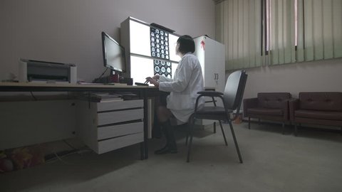 Middle aged female doctor specialist in white gown sits at computer and type on the keyboard medical report after x-ray examination on the light board, steady cam shot, real scene.