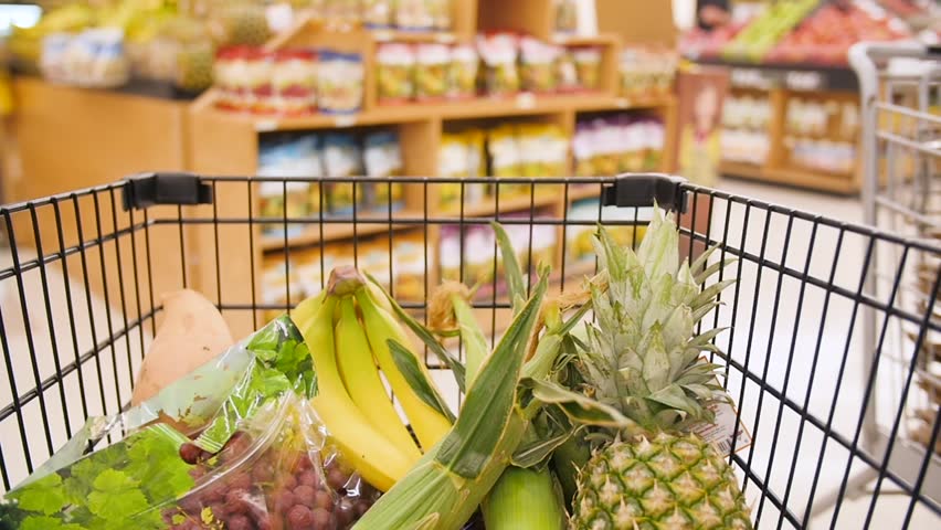 This is a clip of a moving supermarket cart, half-filled with fruits like bananas, pineapple, and grapes. You can see other people in the supermarket at the cart moves through aisles.