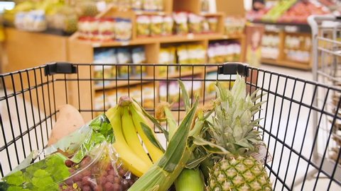 This is a clip of a moving supermarket cart, half-filled with fruits like bananas, pineapple, and grapes. You can see other people in the supermarket at the cart moves through aisles.