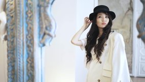 Asian young woman with long hair wearing a black hat is trying on a white coat in a shop. Handheld real time medium shot