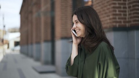 Side view of a lovely young woman wearing a green shirt is talking on the phone in the street standing near a brick building and smiling. Handheld real time medium shot