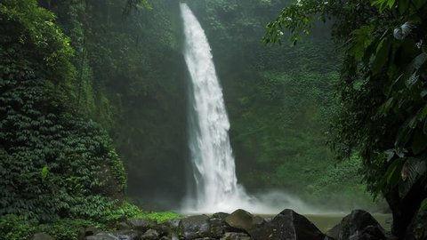 Amazing Nungnung waterfall, reverse slow motion. Some huge rocks seeable in front of frame. Lush green leafes is moving from the breeze, Bali, Indonesia