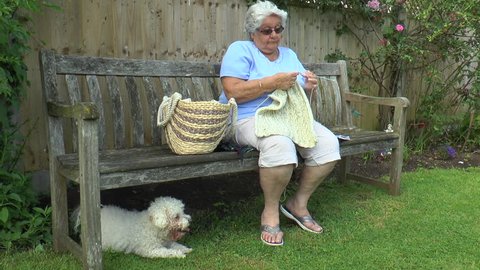 An old woman sitting on a bench, knitting in the shade, with a Bichon Frise dog beneath.