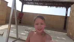 Cute smiling white child cheerfully slides down colorfiul bright waterslide in pool on sunny summer day. Little happy boy holds camera in hand. Real time full hd video footage.