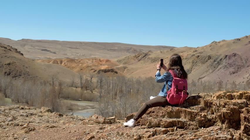Brunette 20s girl in sunglasses, denim jacket and pink backpack makes a photo of a spring canyon with a river, sitting on the rock 4k.