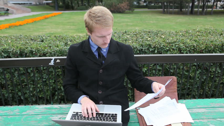 Confident businessman holding document and typing at the laptop in the park